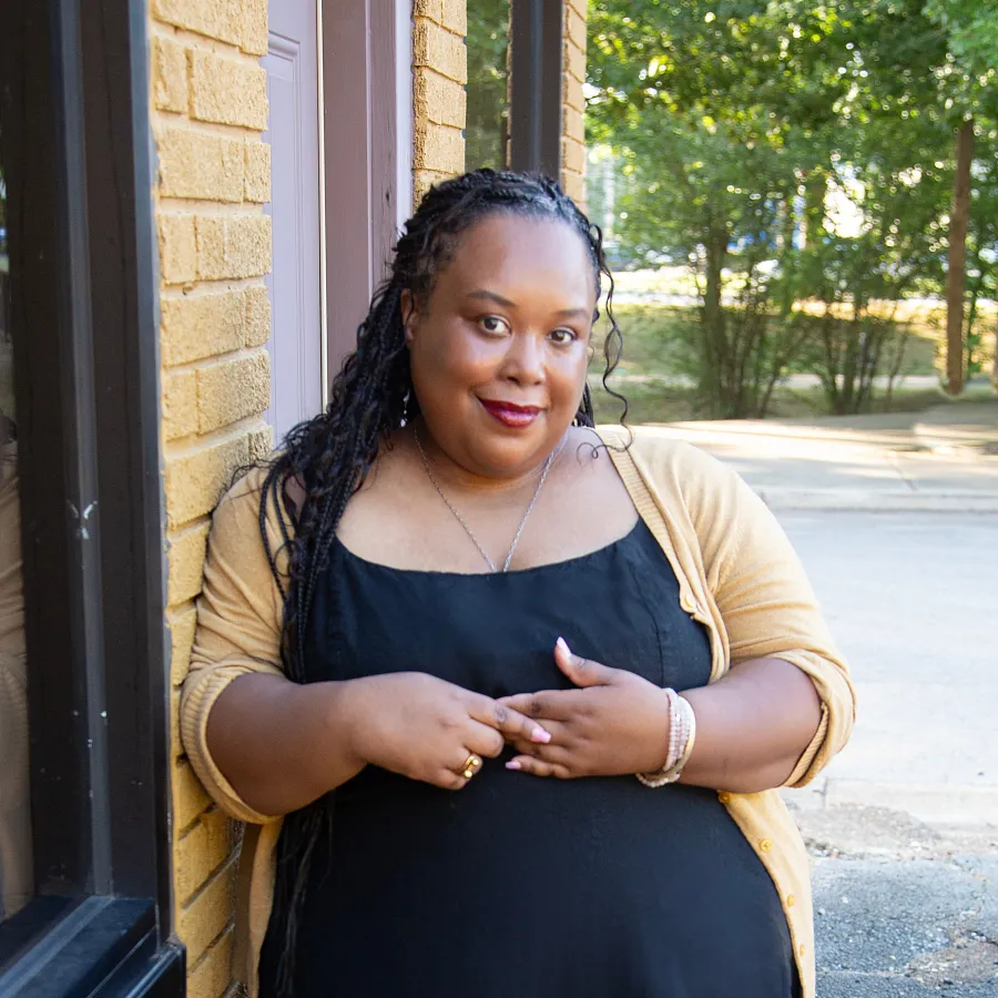 A black woman with braids in a black dress and yellow cardigan smiles at the camera